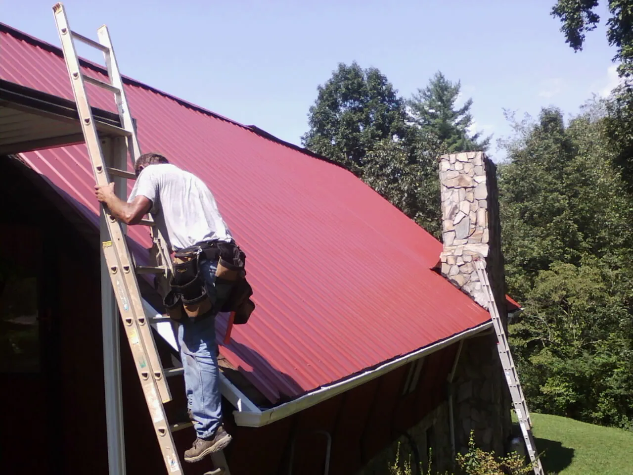 Roofer on a ladder beside a red metal roof and stone chimney — Soffit & Fascia Repair by Roofing Brothers in Nottawa
